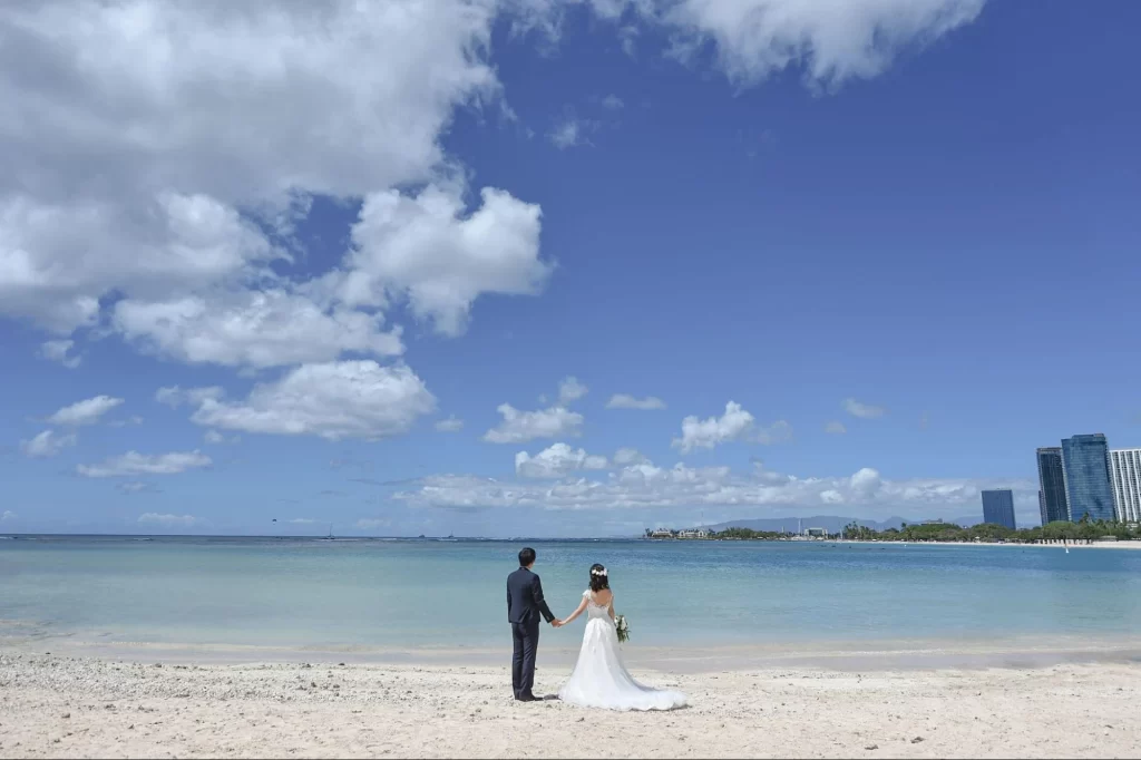 青空の下、ビーチで手をつないで海を眺める新郎新婦の後ろ姿