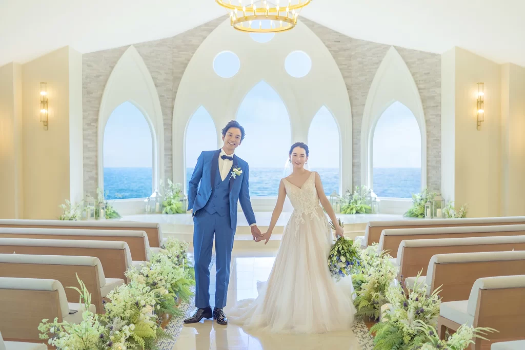 大きなアーチ窓から海が広がるチャペルで、手をつなぐ新郎新婦