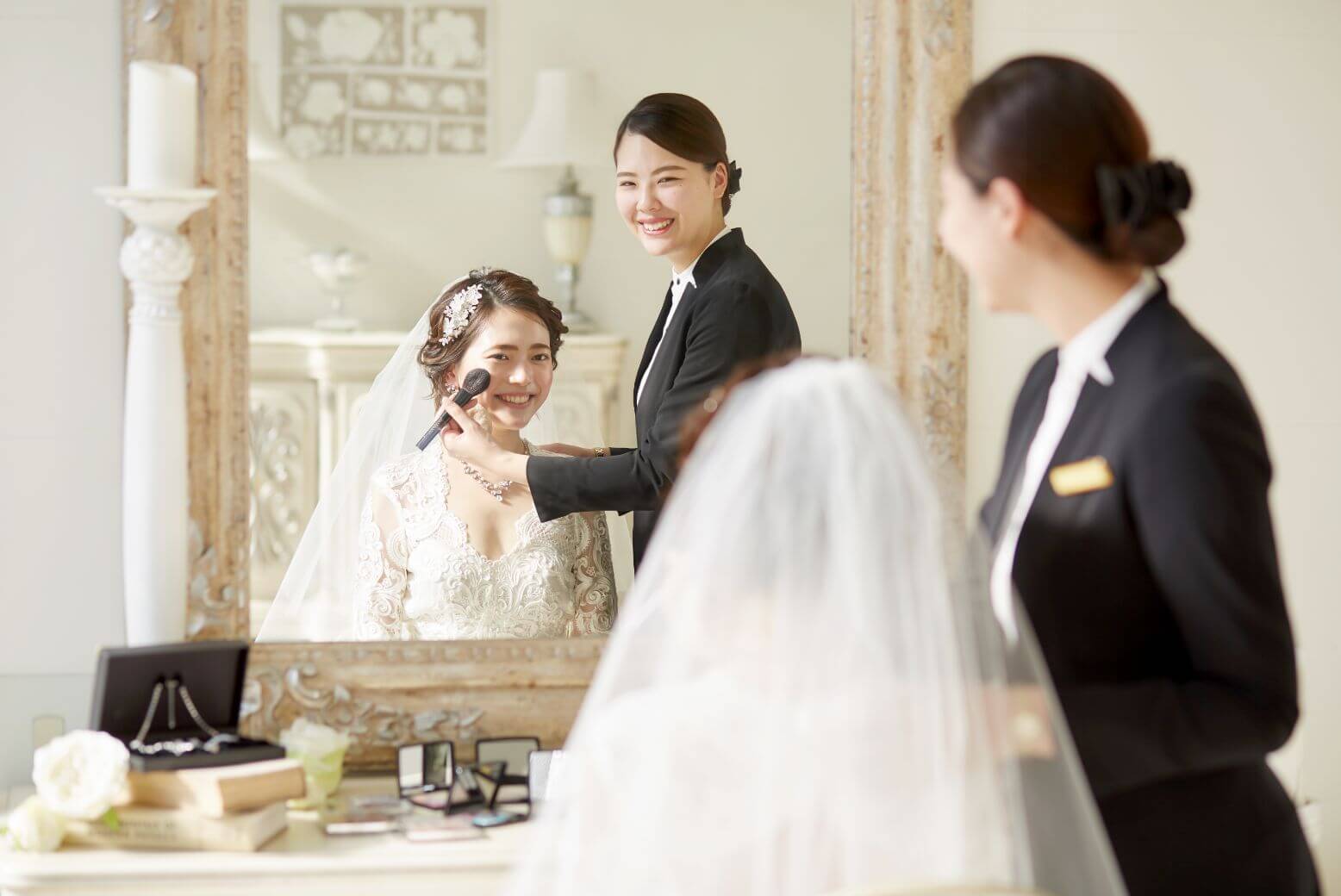 写真：鏡に映るウェディングドレス姿の花嫁とメイクをするヘアメイク担当者の様子
