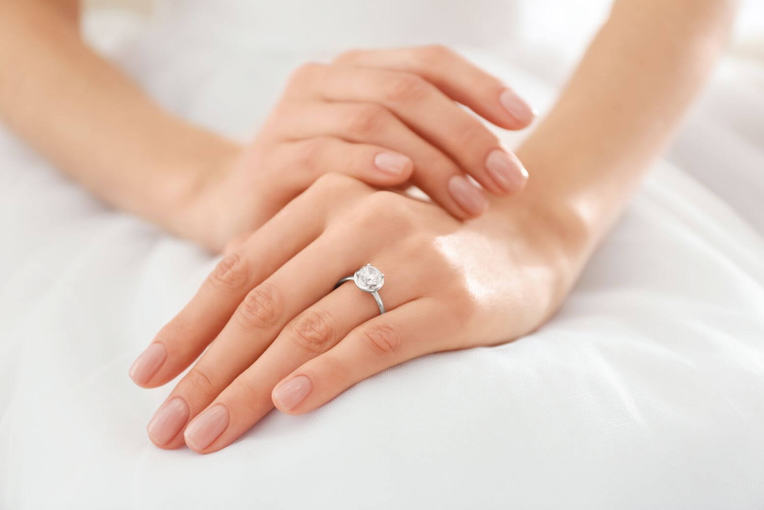 写真：ウェディングドレスの膝の上に重ねられた花嫁の手。左手薬指には大粒ダイヤの婚約指輪が光る