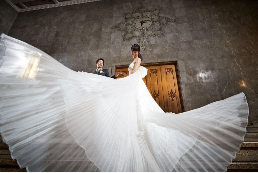 写真:大きく広がるドレープが印象的なウェディングドレスの新婦と、その手を取り見つめる新郎