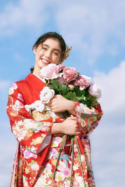 青空のもと、ピンクの花束をかかえて微笑む赤い振袖姿の女性の写真